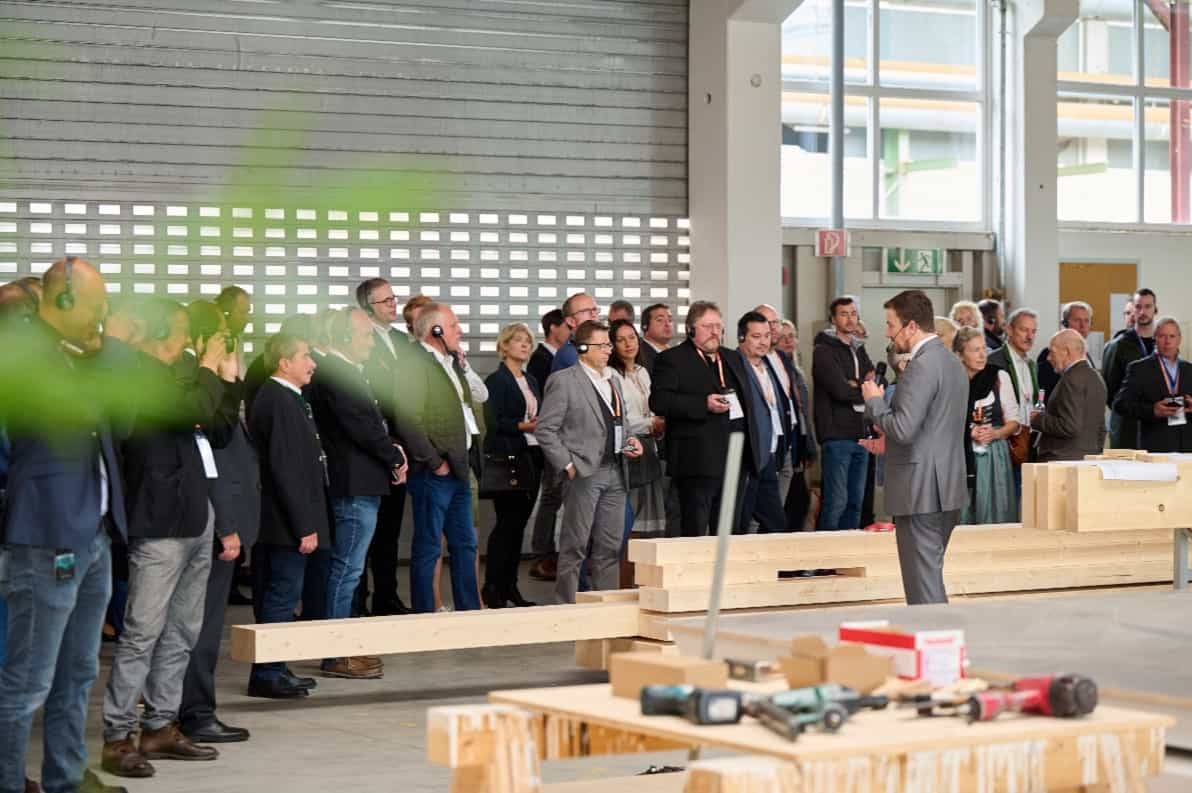 Versammlung von Personen in formeller Kleidung bei einer Präsentation in einer Werks- oder Fabrikhalle. Sprecher hält Mikrofon.