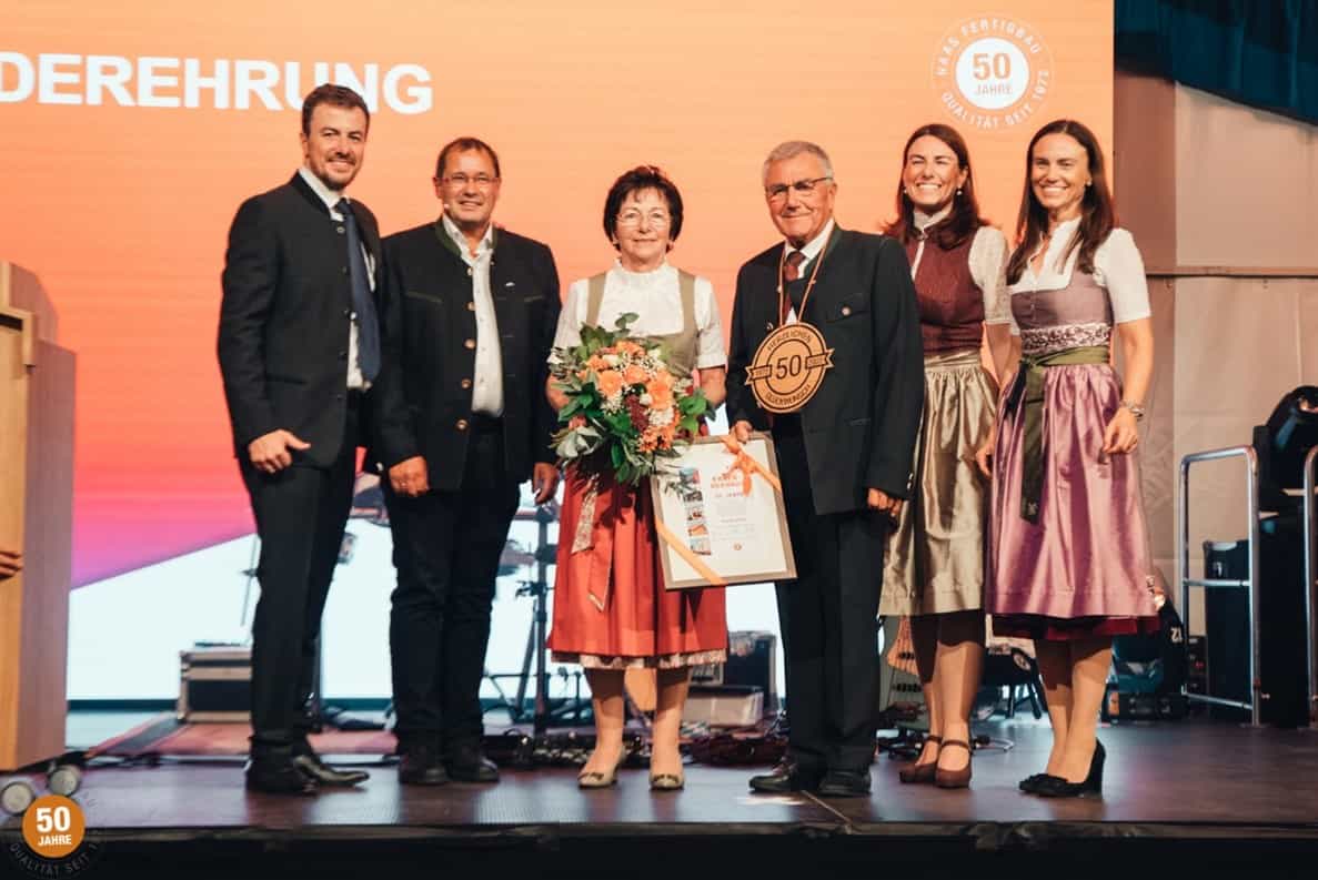 Sechs Personen in festlicher Kleidung auf einer Bühne, zwei halten Blumen und eine Auszeichnung für 50 Jahre, Hintergrund mit Jubiläumslogo.