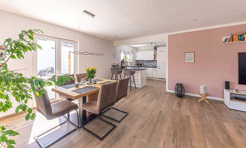 Offener Wohn-Essbereich mit Holztisch, Küche und roséfarbener Wand im Doppelhaus Haas D 137 B