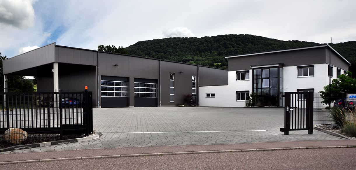 Produktionshalle mit Bürogebäude der APA-Tec GmbH mit grauer Fassade, Vordach und gepflastertem Hof