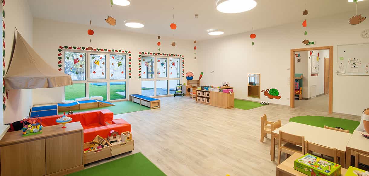 Gruppenraum der Kindertagesstätte Groß-Enzersdorf mit rotem Sofa, Holzmöbeln und runden Deckenleuchten