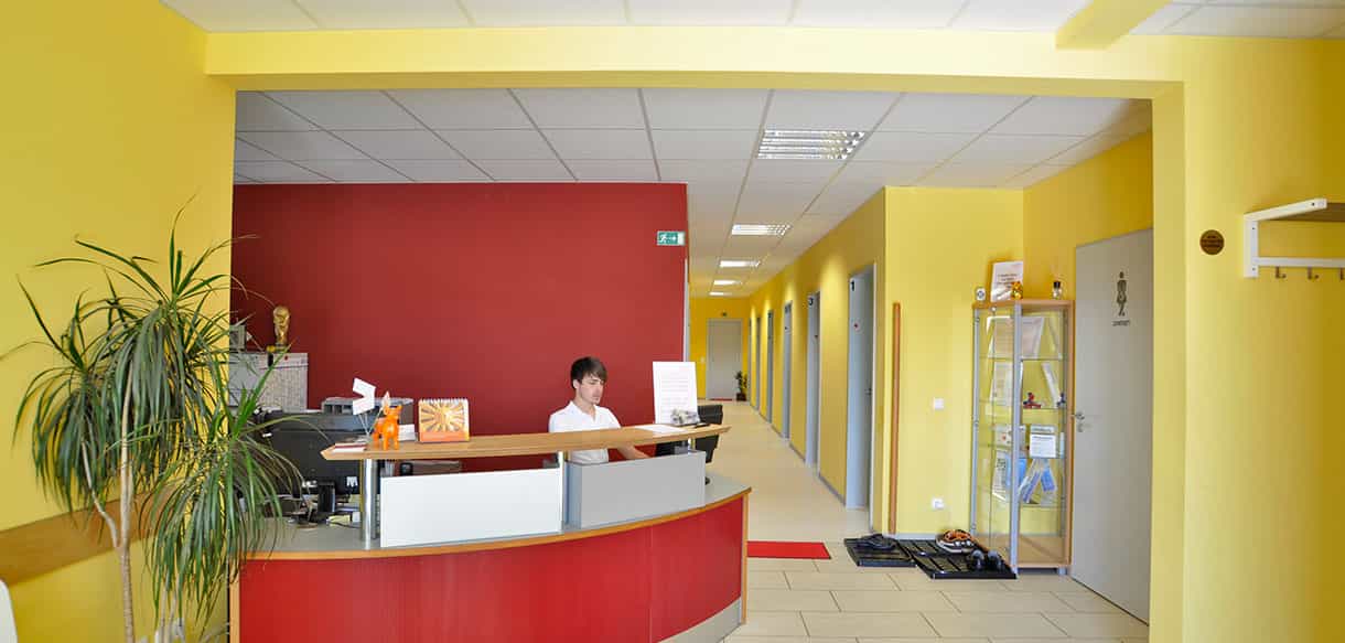 Empfangsbereich der Arcus Physiotherapiepraxis mit roter Theke und gelben Wänden, Mitarbeiterin am Empfang