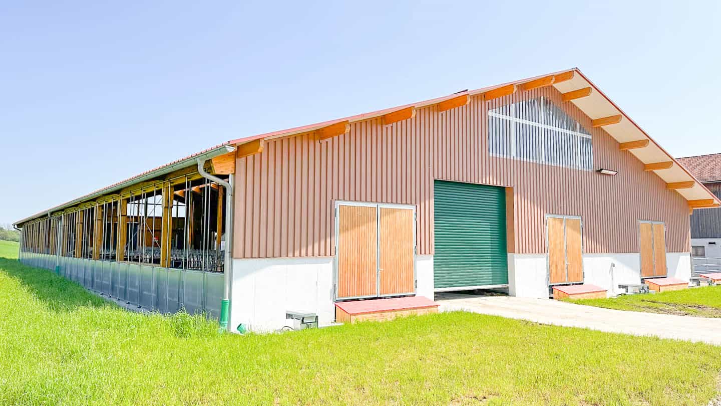 Milchviehlaufstall in Frauenneuharting mit Holzfassade, grünem Rolltor und Windschutznetz