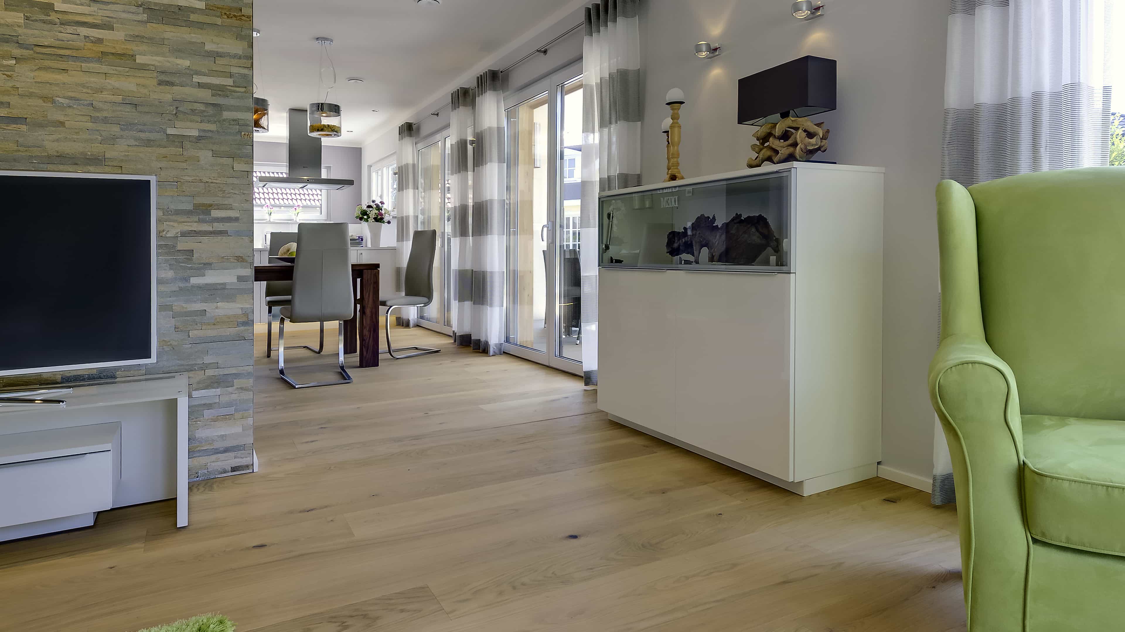 Wohnzimmer mit Natursteinwand, Fernseher und grünem Ohrensessel im Musterhaus Haas MH Mannheim J 159