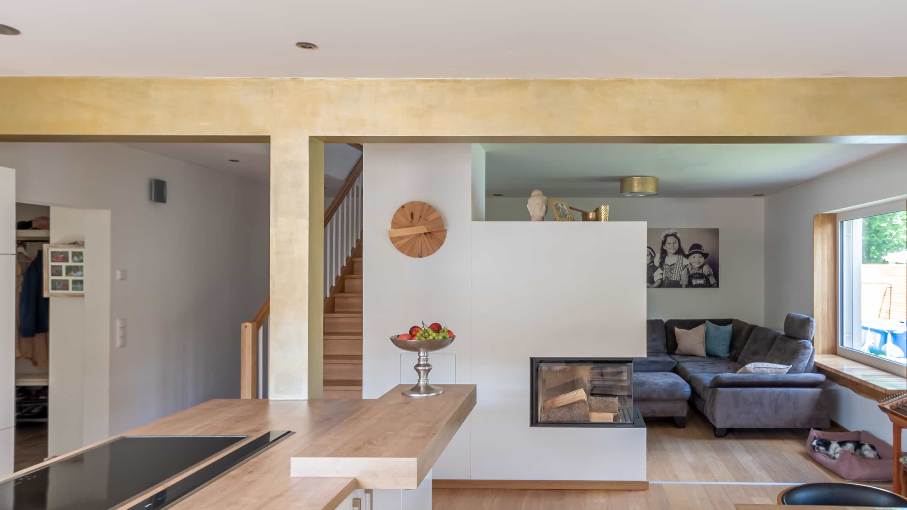 Moderne offene Wohnküche mit Holzfußboden, Kücheninsel und Kamin; dahinter ein Wohnzimmer mit grauer Couch und großen Fenstern.