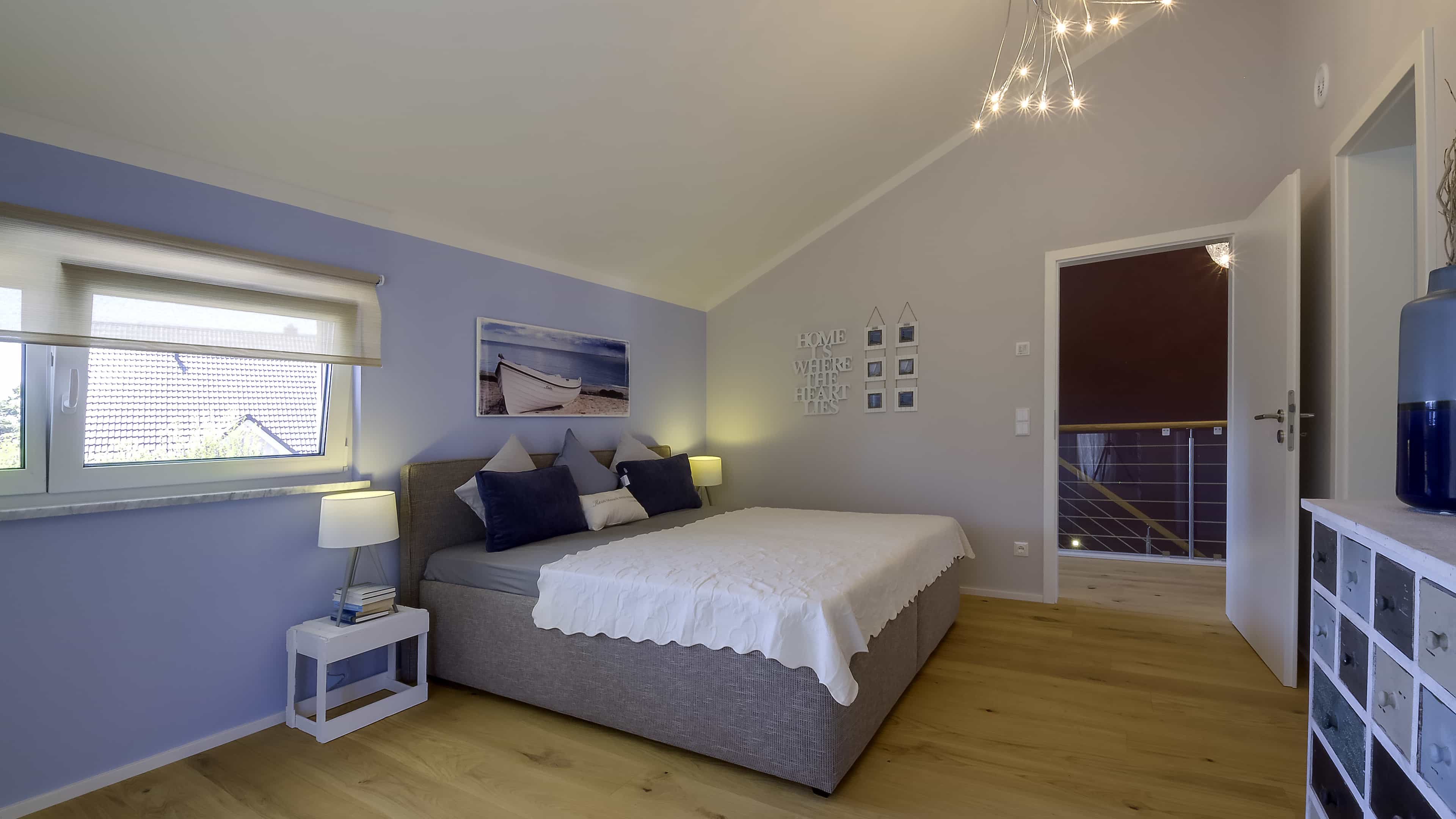 Schlafzimmer mit grauem Bett und fliederfarbener Wand unter der Dachschräge im Haas Musterhaus Mannheim J 159