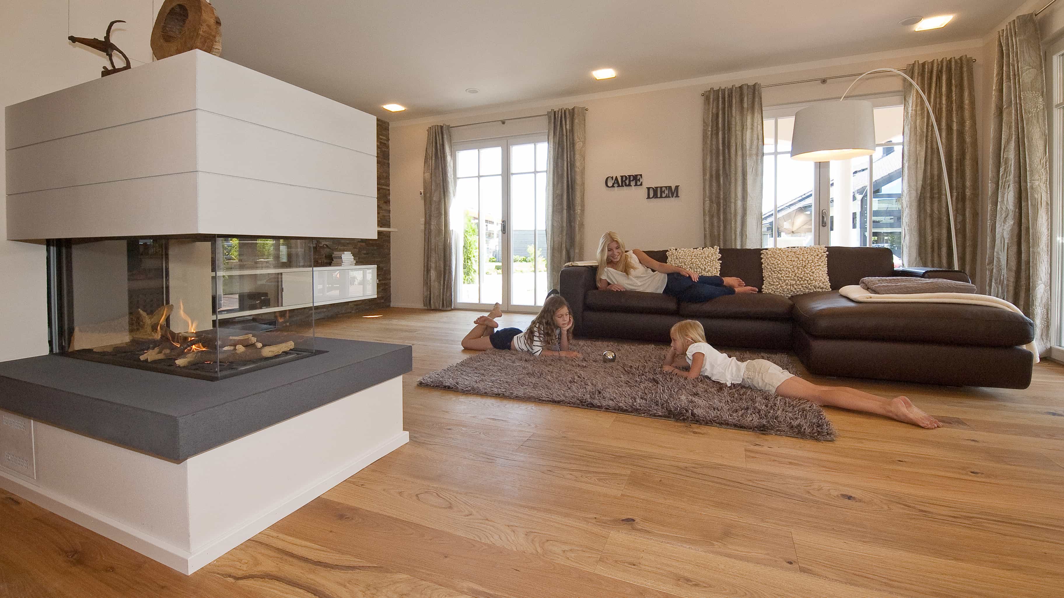 Wohnzimmer mit Kamin, brauner Ledercouch und Kindern auf dem Teppich im Musterhaus Haas MH Poing 187