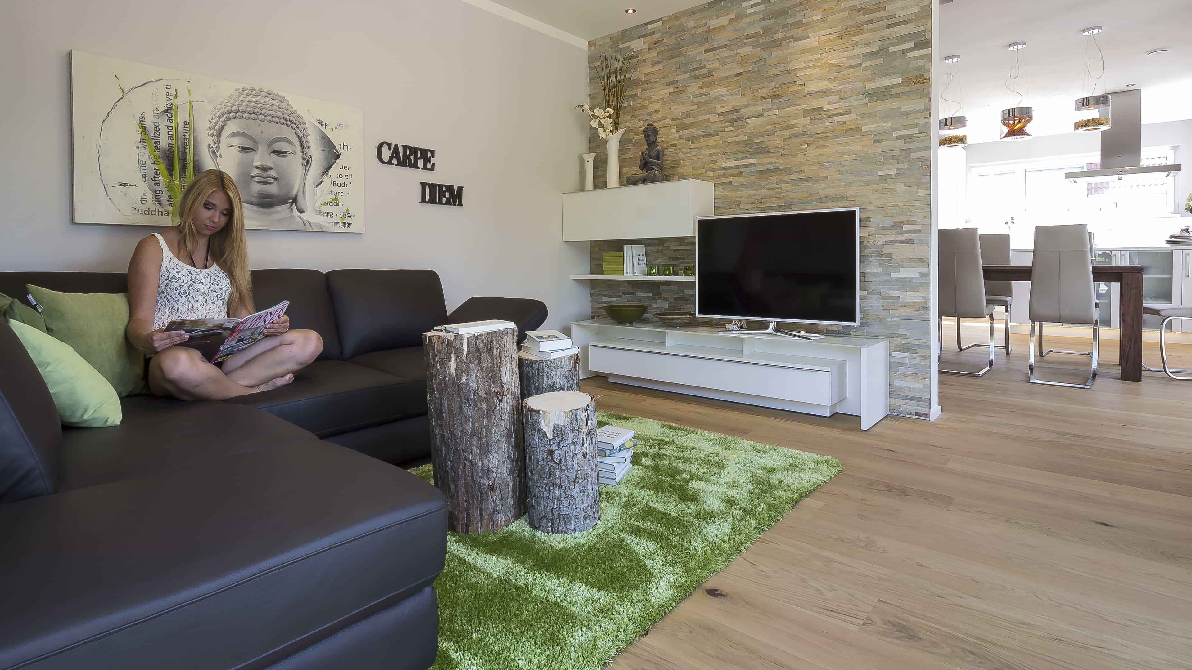 Wohnzimmer mit Sofa, Natursteinwand und Fernseher im Musterhaus Haas MH Mannheim J 159