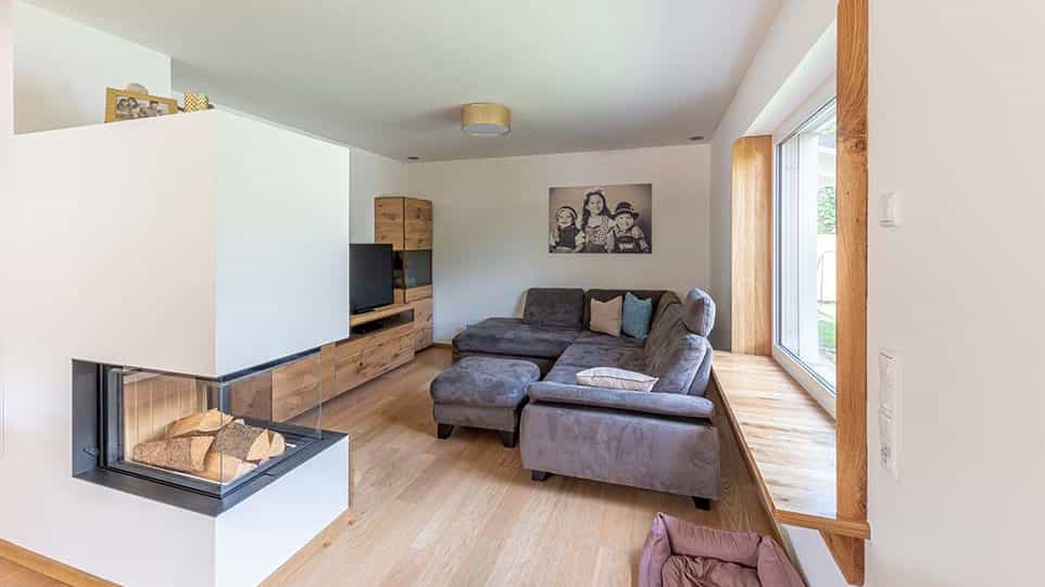 Gemütliches Wohnzimmer mit grauem Ecksofa, modernem Kamin und Holzfußboden, Wandbild im Hintergrund und großem Fenster mit Sitzbank.