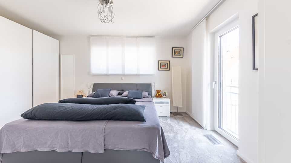 Schlafzimmer mit grauem Bett und weißen Schränken im Fertighaus der Familie Schmid