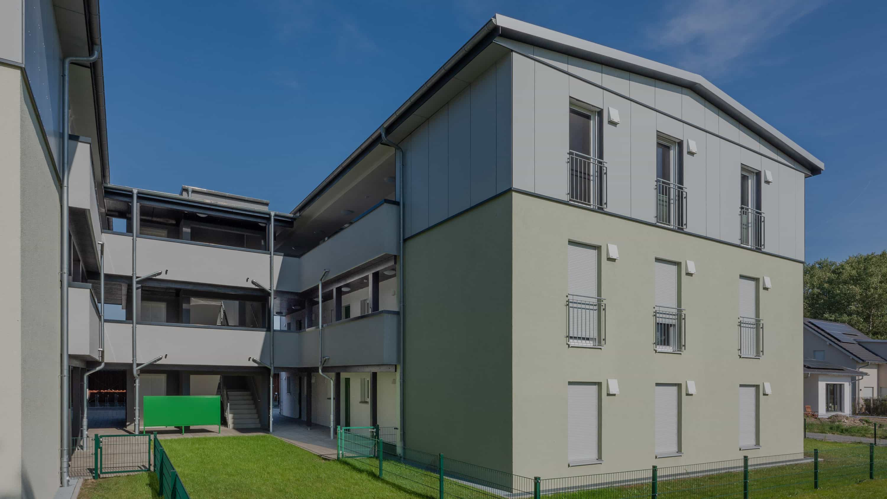 Dreigeschossiges Mehrfamilienhaus mit grüngrauer Fassade und Laubengang im Wohnquartier Zum Glück Bötzow