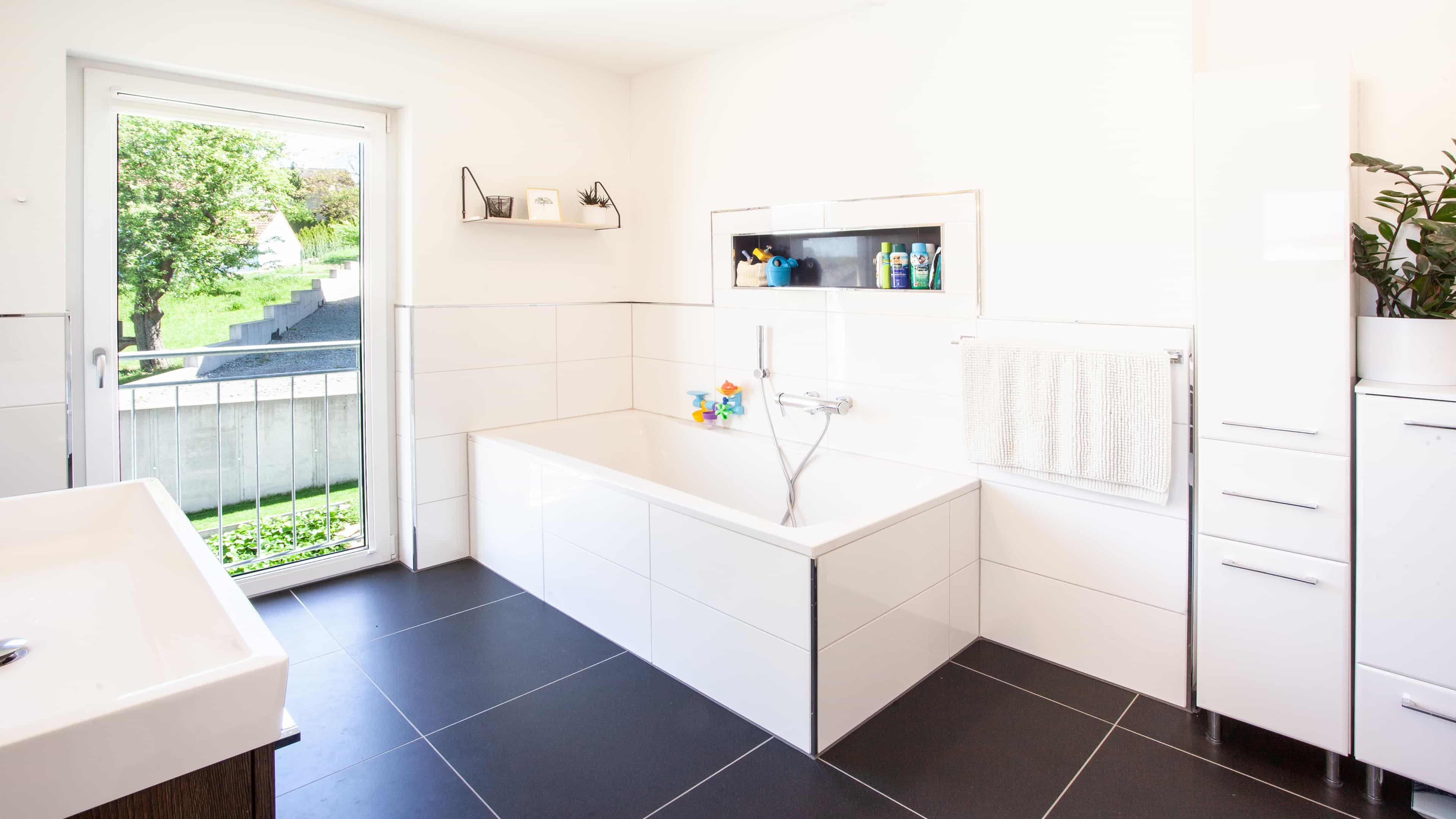 Modernes Badezimmer mit Badewanne, großem Fenster und schwarzen Fliesen; Blick in den Garten.