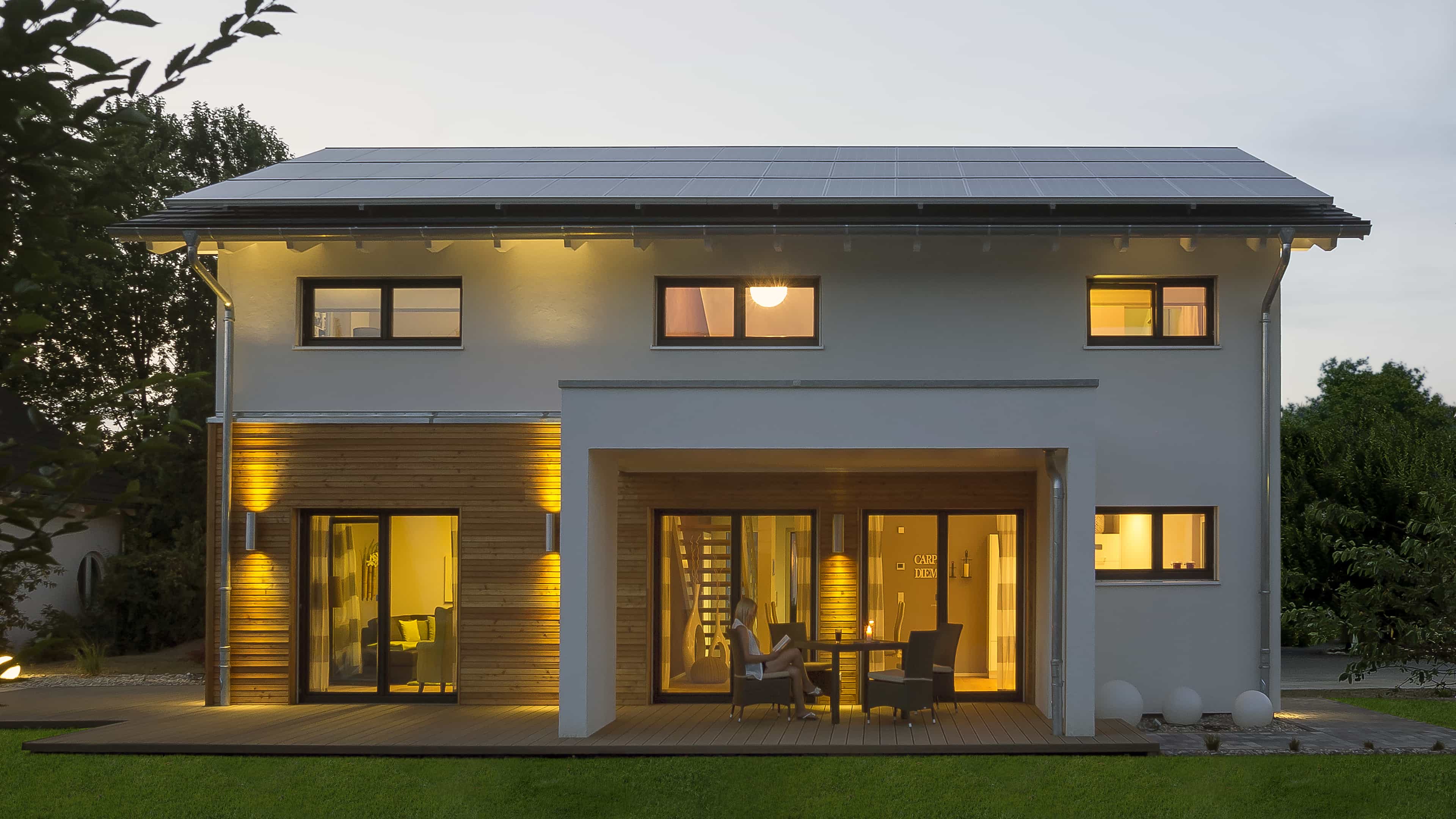 Gartenseite des Musterhauses Haas MH Mannheim J 159 mit beleuchteter Terrasse in der Dämmerung